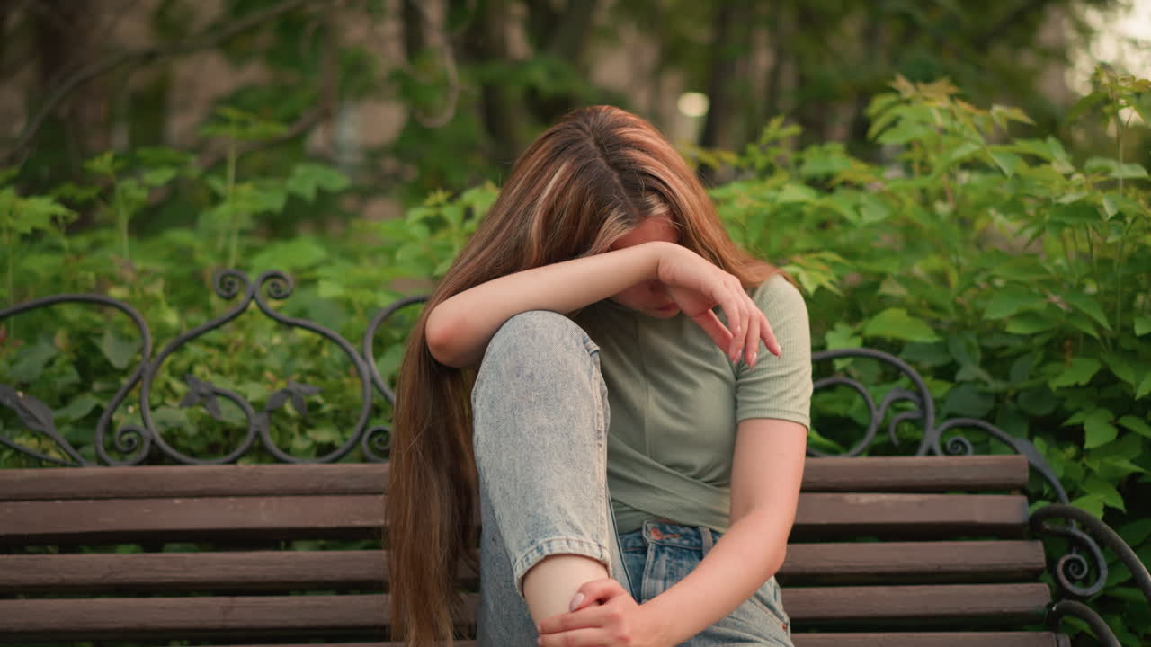 una mujer joven en vaqueros y zapatillas blancas se sienta en un banco de madera en el parque, apoyando su cabeza en su brazo con una expresión reflexiva y sombría, rodeada de exuberante vegetación, árboles y edificios en el fondo