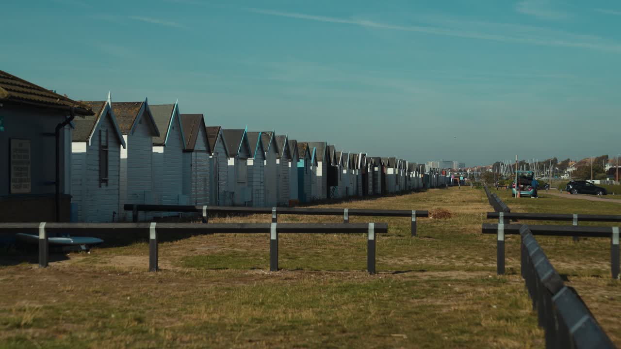 Un'ampia ripresa al rallentatore di cabine sulla spiaggia inglese con uno stormo di uccelli che volano via. Cielo azzurro e spiaggia tranquilla.
