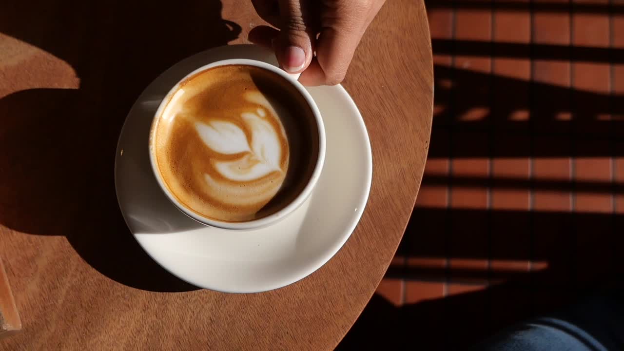 Mano che prende una tazza di caffè latte art da un tavolo di legno sotto la luce del sole