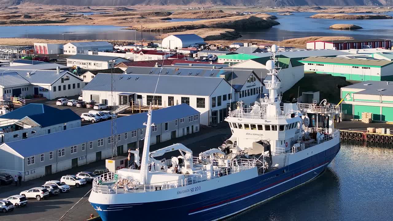 The amazing drone footage captures the picturesque coastal scenery of Hofn, showcasing the sea, boats, hills, and the cityscape of the City and Harbor of Hofn in Iceland