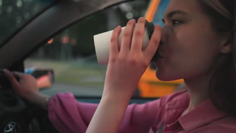 woman driving while holding coffee cup in one hand and steering the wheel with the other hand, a blurred view of an orange truck passes by on the road, with a building in the background