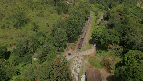 Pullback-Aerial-Drone-Shot-of-Train-Departing-Demodara-Train-Station-Looking-Down-Valley-on-Sunny-Day-in-Sri-Lanka