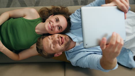 funny couple taking selfie laying couch top view. happy man photographing tablet