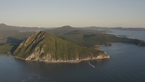 a landscape view of a pyramid-shaped mountain located at a river estuary flowing into the sea, with green vegetation on its sides, mountain ridge in the background, on the sunset