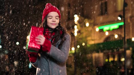 retrato de una linda chica feliz sosteniendo un regalo de navidad y sonriendo a la cámara en la calle mientras nieva en navidad