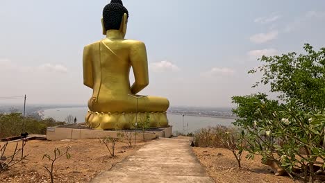 camera steadily moves forward along a dirt path toward a massive golden buddha statue, set on a hilltop with river and sky in background, under bright daylight