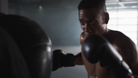 hombre afroamericano con guantes de boxeo entrenando en un edificio vacío golpeando una bolsa de boxeo