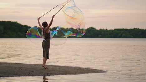 a young female artist shows a soap bubble show blowing up huge soap bubbles on the shore of a lake at sunset. show a beautiful show of soap bubbles in slow motion