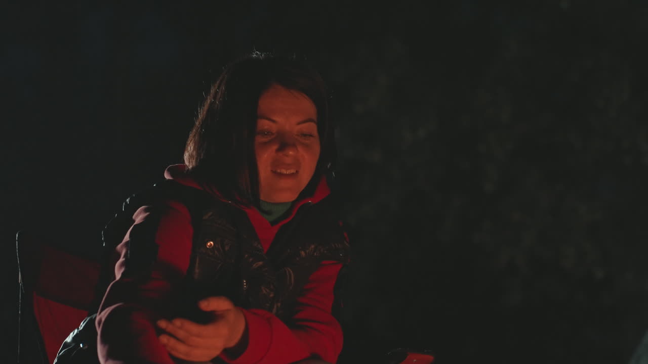 Primo piano di donna bianca accanto al fuoco da campo, giacca rossa e bastone. Primo piano notturno mostra il bagliore sul viso, braci calde, scintille delicate e fumo di carbone. Campeggiatrice solitaria che si scalda le mani, attizza le braci, sorridendo in silenzio.