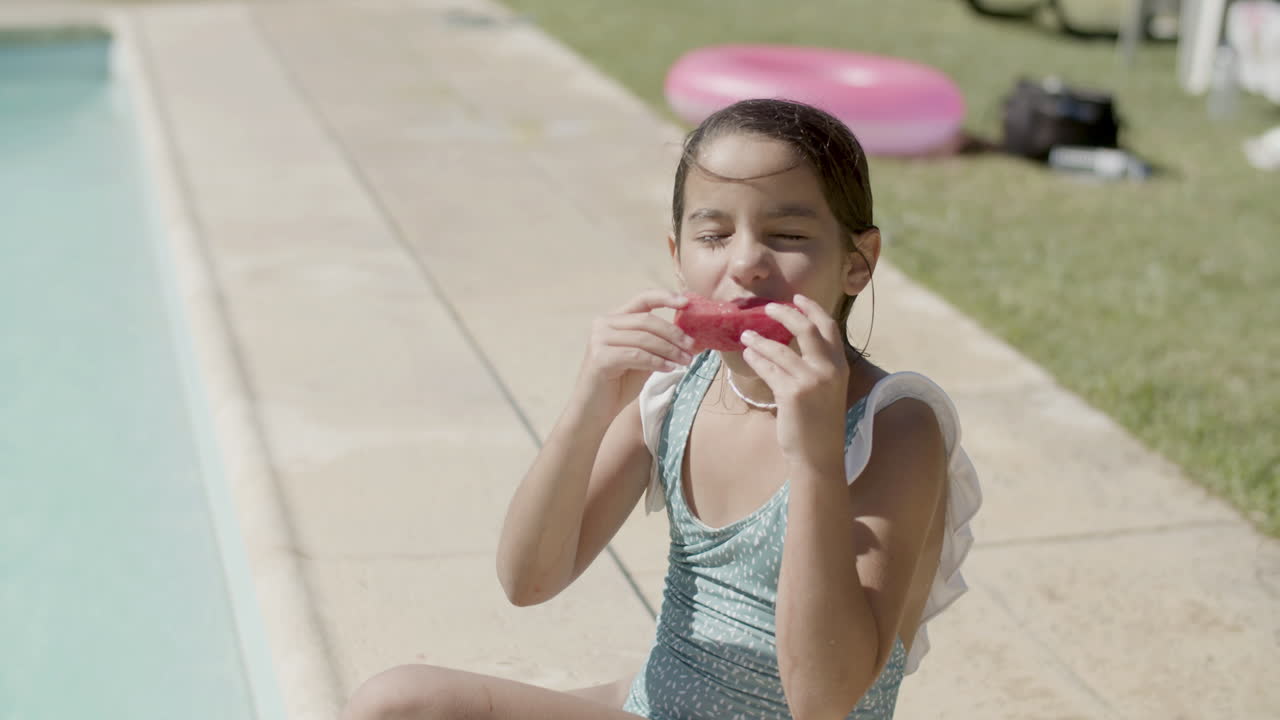 una chica linda feliz sentada a la orilla de la piscina mordiendo una sandía jugosa.