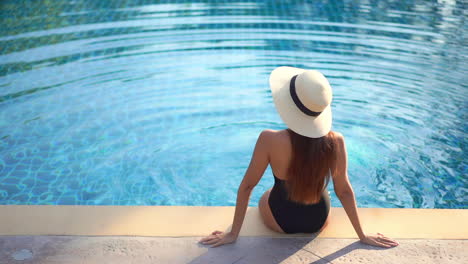 sitting on the edge of a pool, a young woman with her back to the camera, gently kicks her feet creating ripples in the water and time