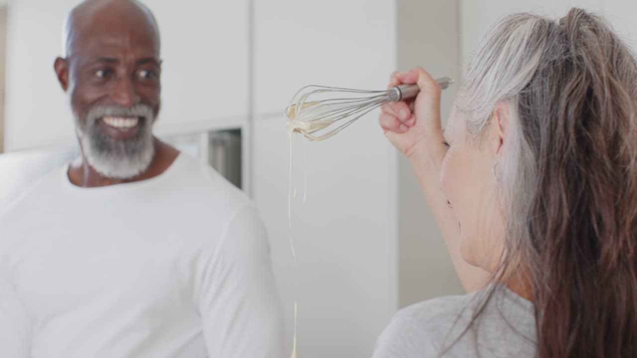 Coppia multietnica che sorride, la donna solleva una frusta mostrando l'impasto nella ciotola, mentre l'uomo si gira al bancone della cucina.
