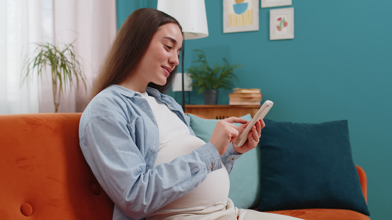 스마트폰을 사용하는 미소 짓는 임신한 여성이 가정 소파에 앉아있는 아기를 위해 온라인 구매를합니다.