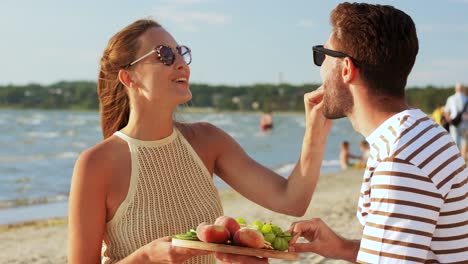 Pareja-Feliz-Con-Comida-Haciendo-Un-Picnic-En-La-Playa.-Concepto-De-Ocio,-Relaciones-Y-Personas.-Pareja-Feliz-Con-Comida-Comiendo-Uvas-Y-Haciendo-Un-Picnic-En-La-Playa.