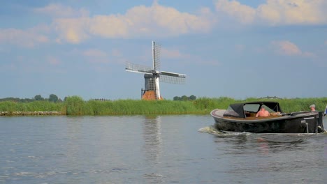 Antiguo-Molino-De-Viento-Holandés-En-Un-Dique-Cerca-Del-Agua,-Un-Pequeño-Carro-Rodado-En-Cámara-Lenta-Y-Otro-Barco-Navega-De-Derecha-A-Izquierda