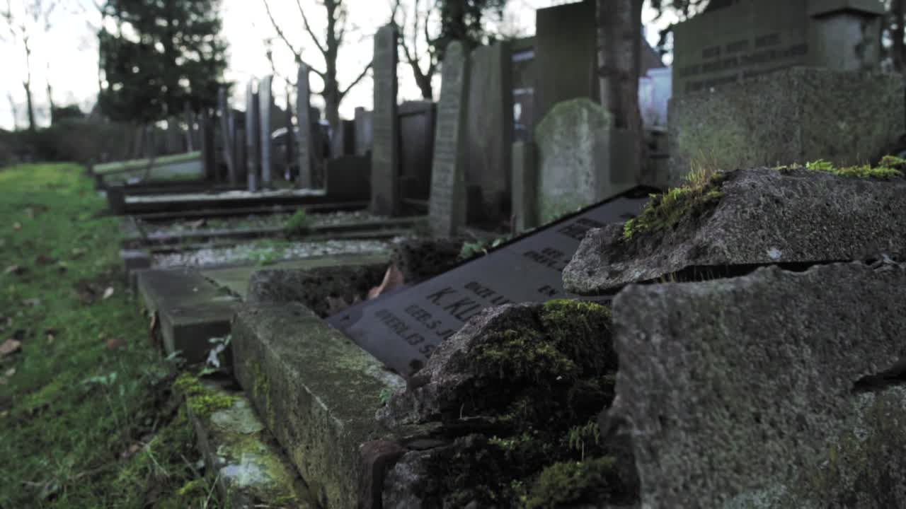 Atmospheric footage of an old cemetery in Texel, Netherlands, showing weathered and moss-covered gravestones under a cloudy sky, evoking history, decay, and the passage of time