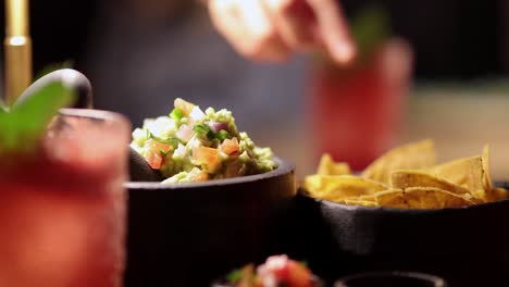 hands dip tortilla chips into fresh guacamole at a dimly lit bangkok restaurant. a vibrant cocktail sits nearby, enhancing the lively atmosphere