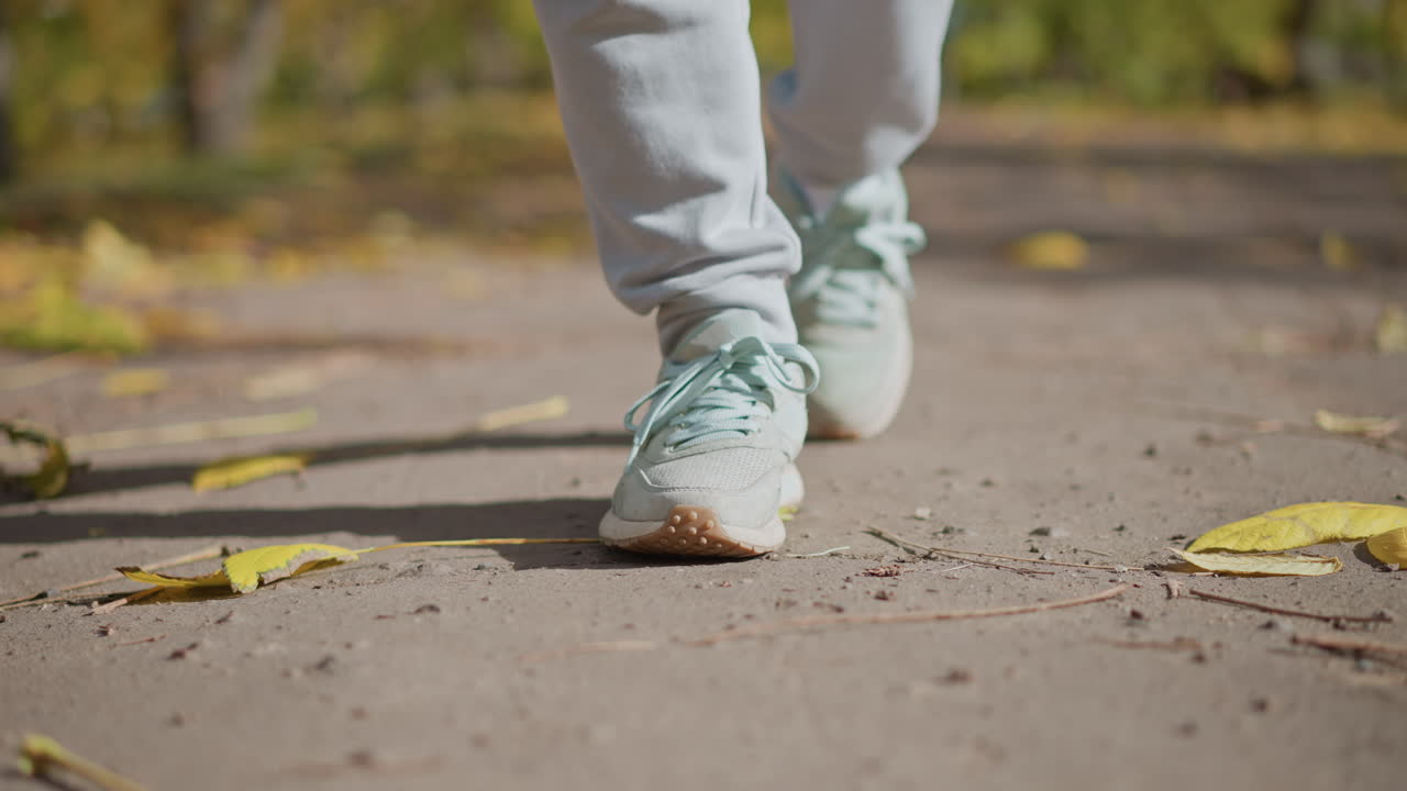 close up of person walking along autumn path strewn with fallen yellow leaves wearing light jogger pants and pastel sneakers, showing heel lift and cast shadow on pavement with blurred foliage