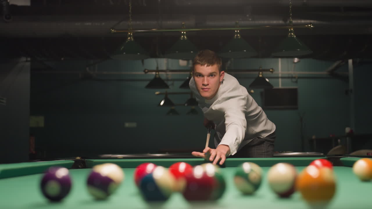 Close-up van een biljarter in een wit overhemd en een grijze broek die met intense focus een keu vasthoudt. Groene pooltafel met verspreide biljartballen. Bovenlichten werpen dramatische schaduwen