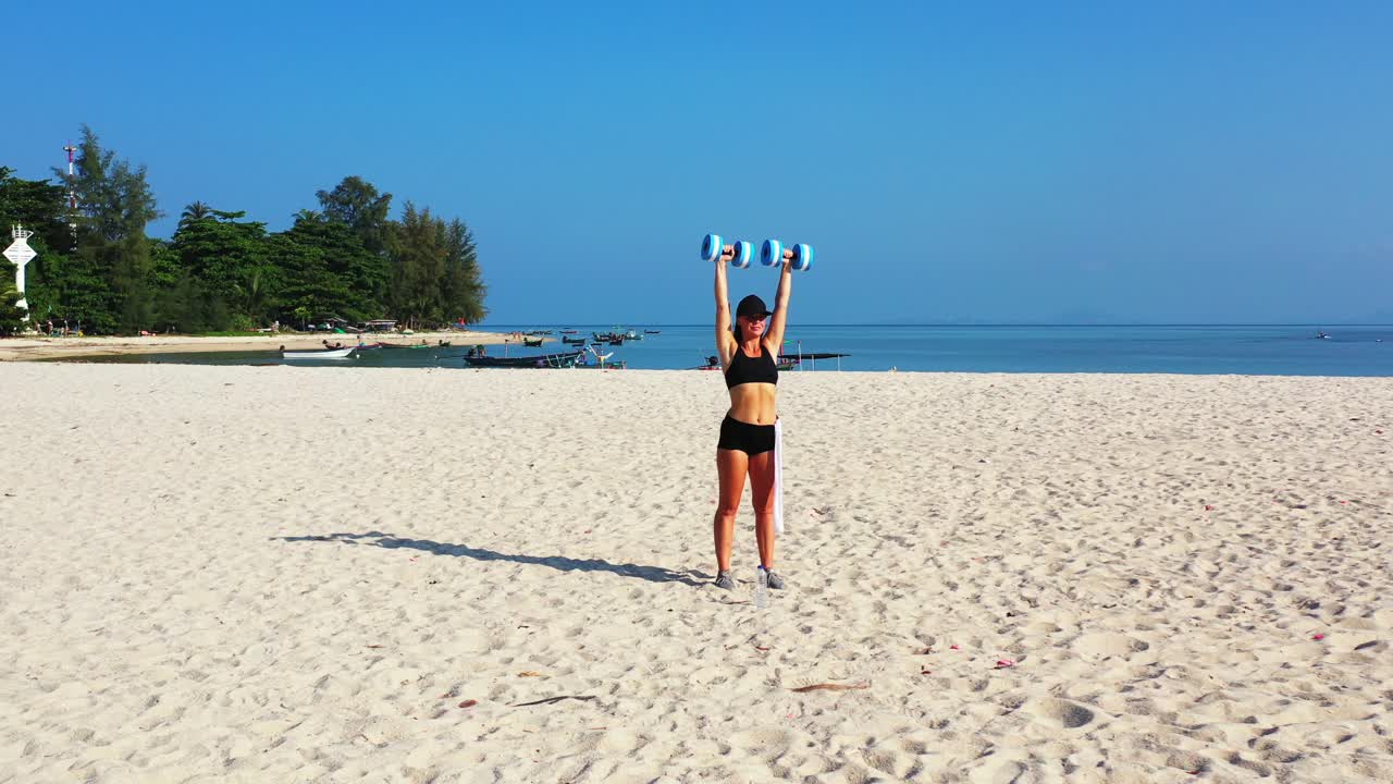 Beautiful girl exercising with two weights on calm exotic beach with wide white sand near blue sea on a sunny day in Hawaii