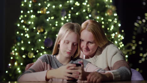 Mom-and-daughter-are-looking-at-the-smaptphone-screen-together.-Sitting-in-front-of-the-blurred-lights-of-the-Christmas-tree