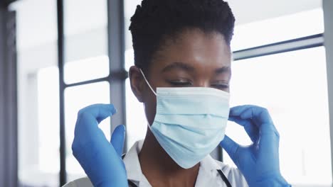 Portrait-of-african-american-female-doctor-wearing-surgical-gloves-putting-on-face-mask