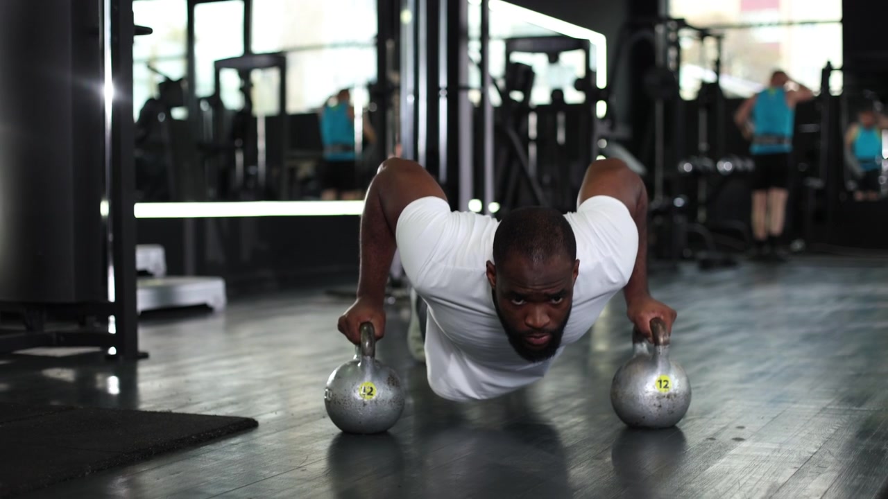Uomo di colore determinato che fa flessioni con kettlebell in una palestra moderna