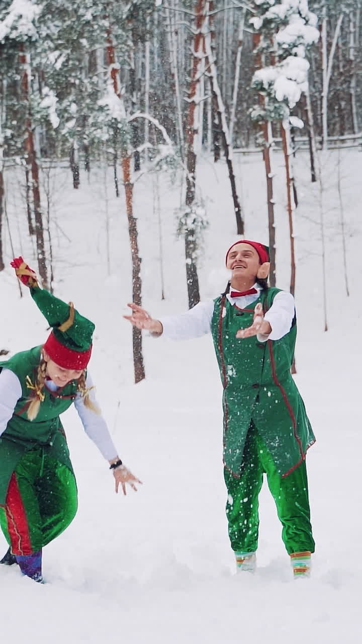 Funny elves in green suits with hats are throwing up snow and looking at it on the background of a winter park. Slow motion Vertical video