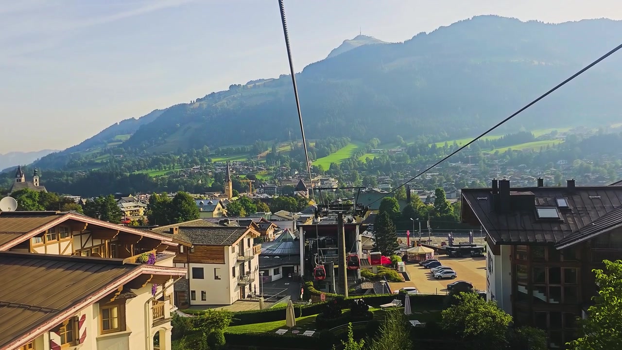 Gondole rosse salgono sul pendio a Kitzbühel, Austria, circondate da pinete, prati alpini, chalet e le pittoresche montagne del paesaggio estivo del Tirolo sotto il sole estivo.