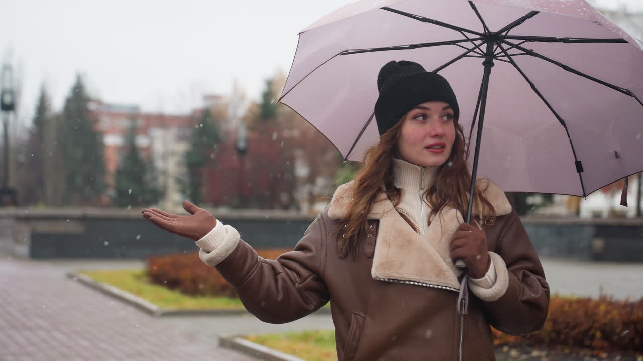 Lady standing under light snowfall, holding an umbrella, and stretching her hand out. She wears a black knit cap and a brown shearling jacket, enjoying winter outdoors with a calm, serene expression