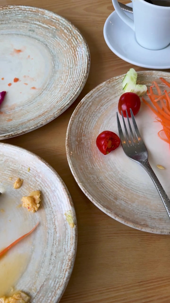 Finished breakfast on a wooden dining table, filmed vertically in an influencer-style POV, showing empty ceramic plates with leftover dragon fruit, scrambled eggs and vegetables.