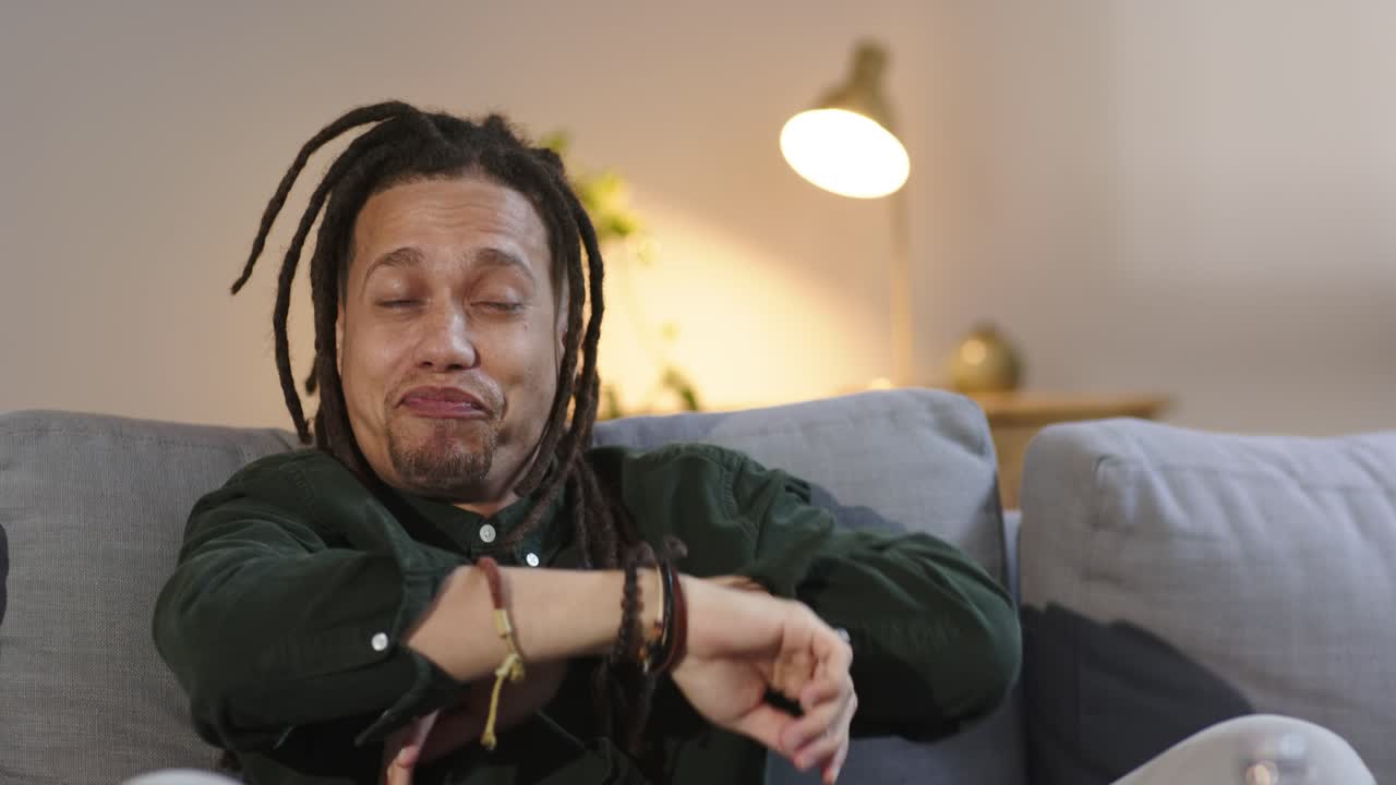 Uomo con dreadlock che indossa una maglietta verde e ride di una battuta fuori campo prima di parlare sul divano di casa