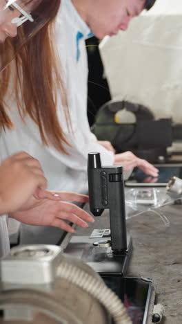 dos técnicos de laboratorio en un taller de automóviles centrados en sus tareas: uno ajusta un microscopio mientras el otro toca una tableta, ambos profundamente absortos en su trabajo