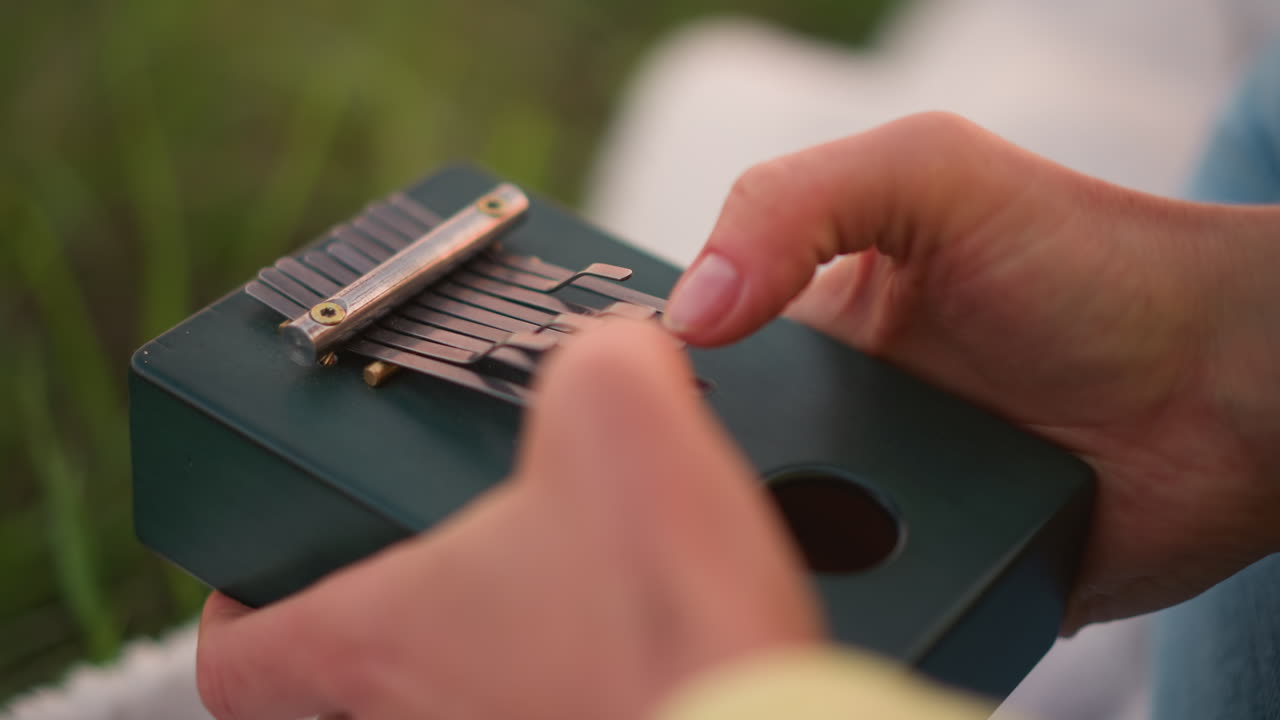white hands composing on green kalimba outdoors, closeup fingers shaping melody over metallic tines, soft grass backdrop and warm glow, focused creative session capturing intimate songwriting