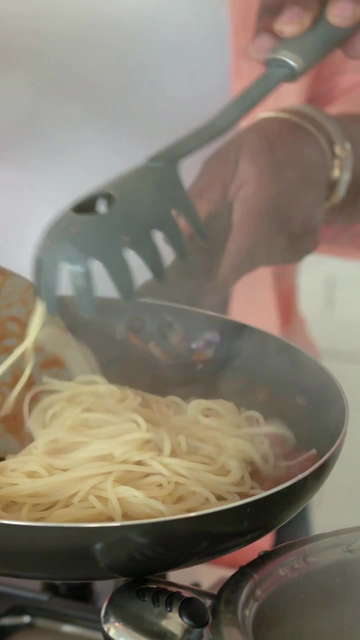 Donna che cucina solleva spaghetti con le pinze, spostando la pasta nel sugo di pomodoro mentre un uomo mescola.