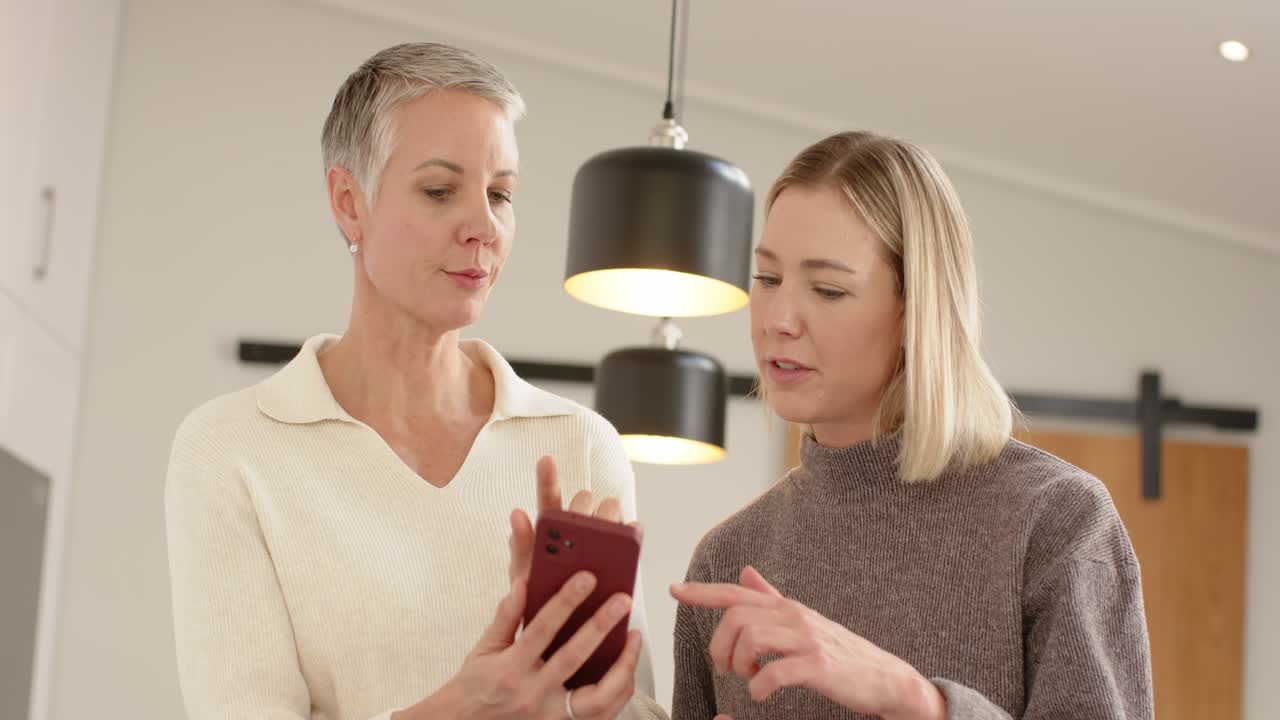 Madre che scorre il suo smartphone rosso mentre la figlia tocca lo schermo con una tazza in cucina.