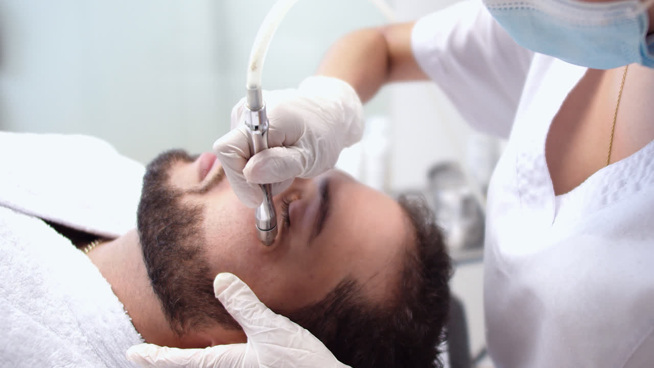 Amazing wide shot of a young man receiving a nanopore beauty treatment from a nurse at a dermatology clinic.