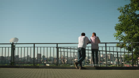 friends seen from behind leaning on iron railing overlooking urban cityscape, lady stands on pavement while boy adjusts his posture and gestures, background features modern buildings