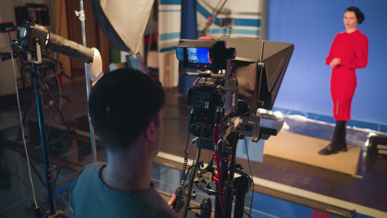 Videographer filming female presenter in red dress on blue screen background inside professional studio with camera monitor, soft lights, and equipment setup visible during production session