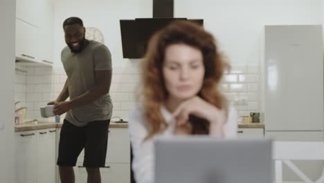 hombre negro feliz llevando tazas de té a una mujer blanca bonita en una cocina moderna.