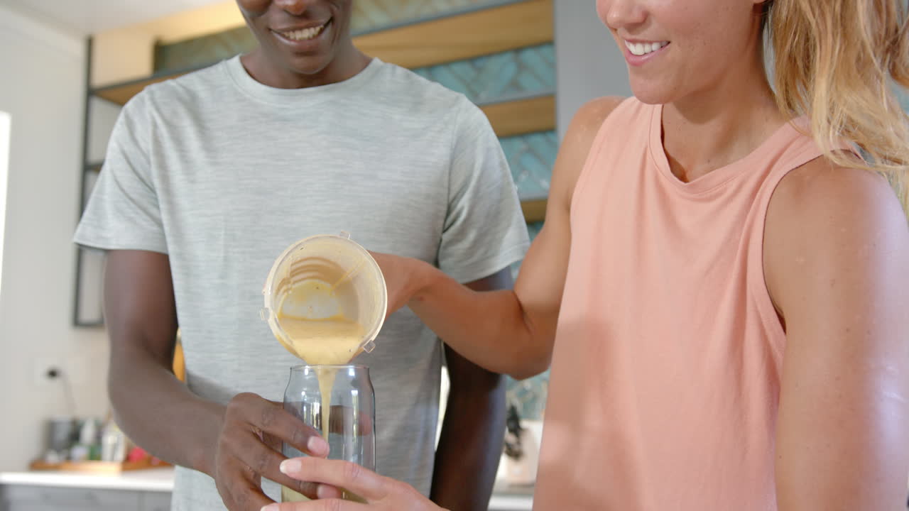 Smiling diverse couple pouring smoothie into glass in modern kitchen together