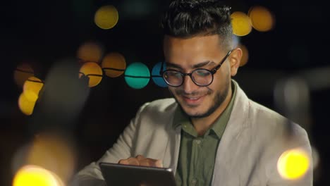smiling arab man playing on digital tablet on rooftop in the night