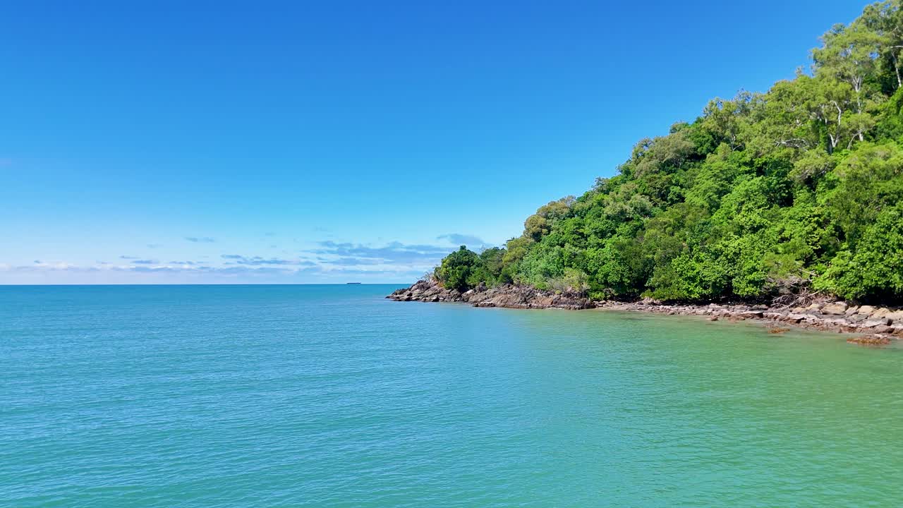 Drone glides past lush rainforest headland, turquoise ocean, and rocky shore under bright daylight