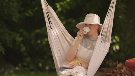 a middle-aged casually looking woman, wearing a striped dress and a sun hat, sitting in the hammock chair, drinking coffee from a small white cup