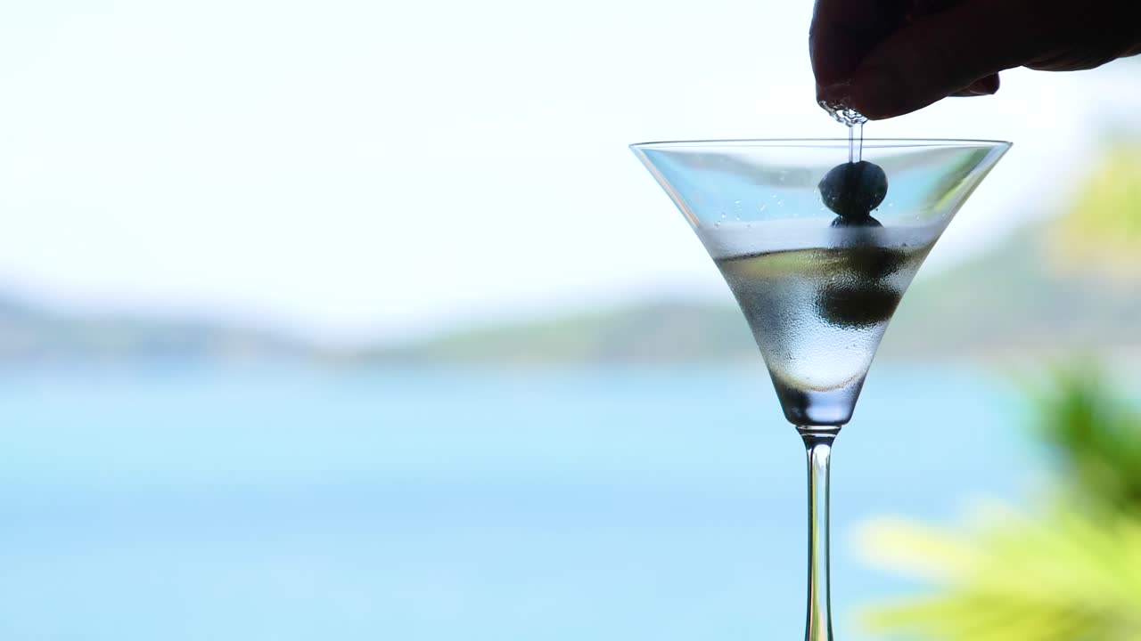 Una mano lascia cadere olive in un bicchiere da martini contro un sereno sfondo marino, catturando un momento tranquillo ed elegante.