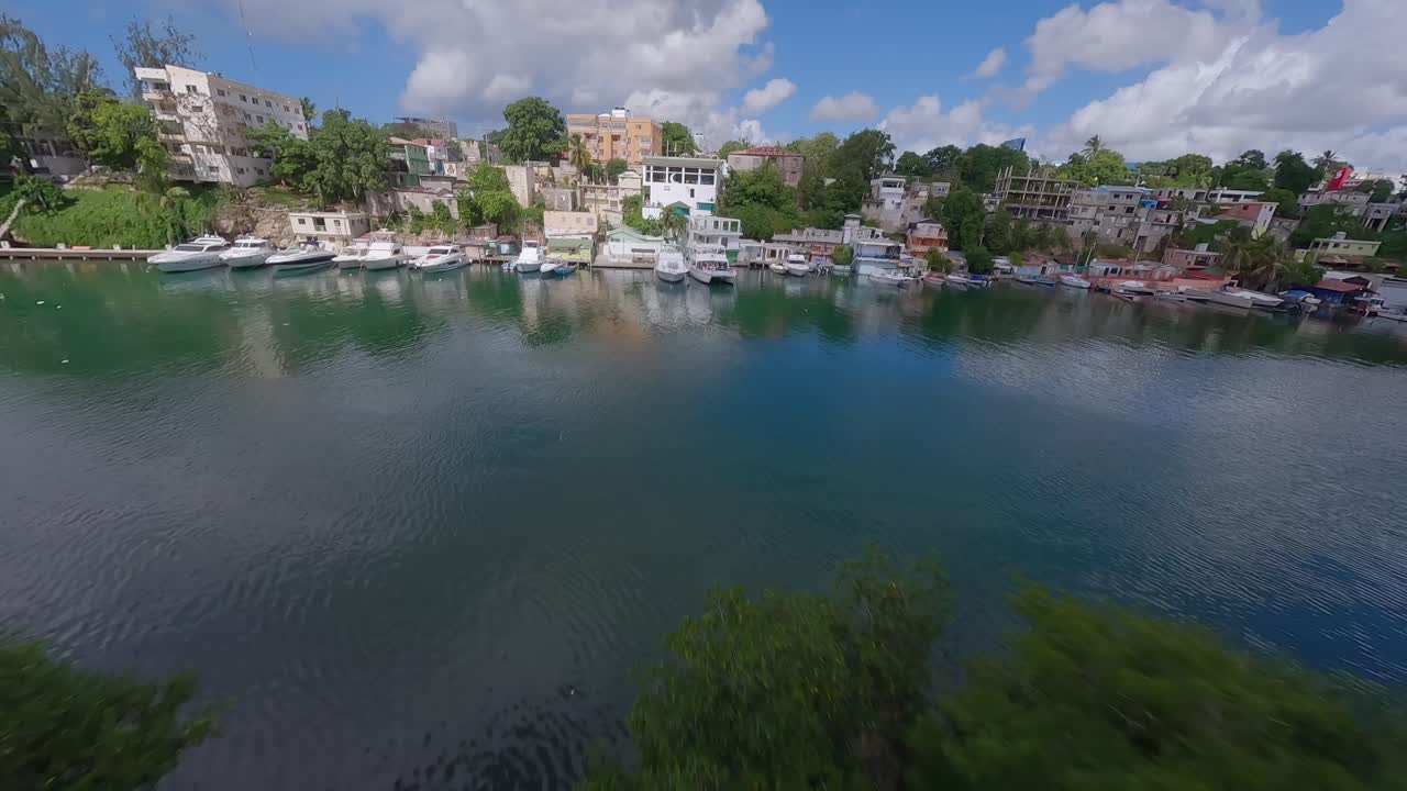 vuelo aéreo de fpv sobre un río tropical con botes de estacionamiento y la ciudad de roma en el fondo, república dominicana