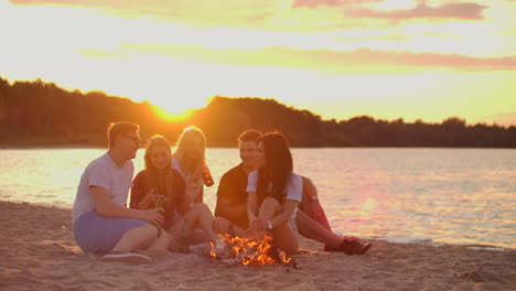 two men and three young girls are sitting around bonfire on the beach with beer. one of the girls is checking her mobile phone and turning music at sunset in summer evening on the lake coast.