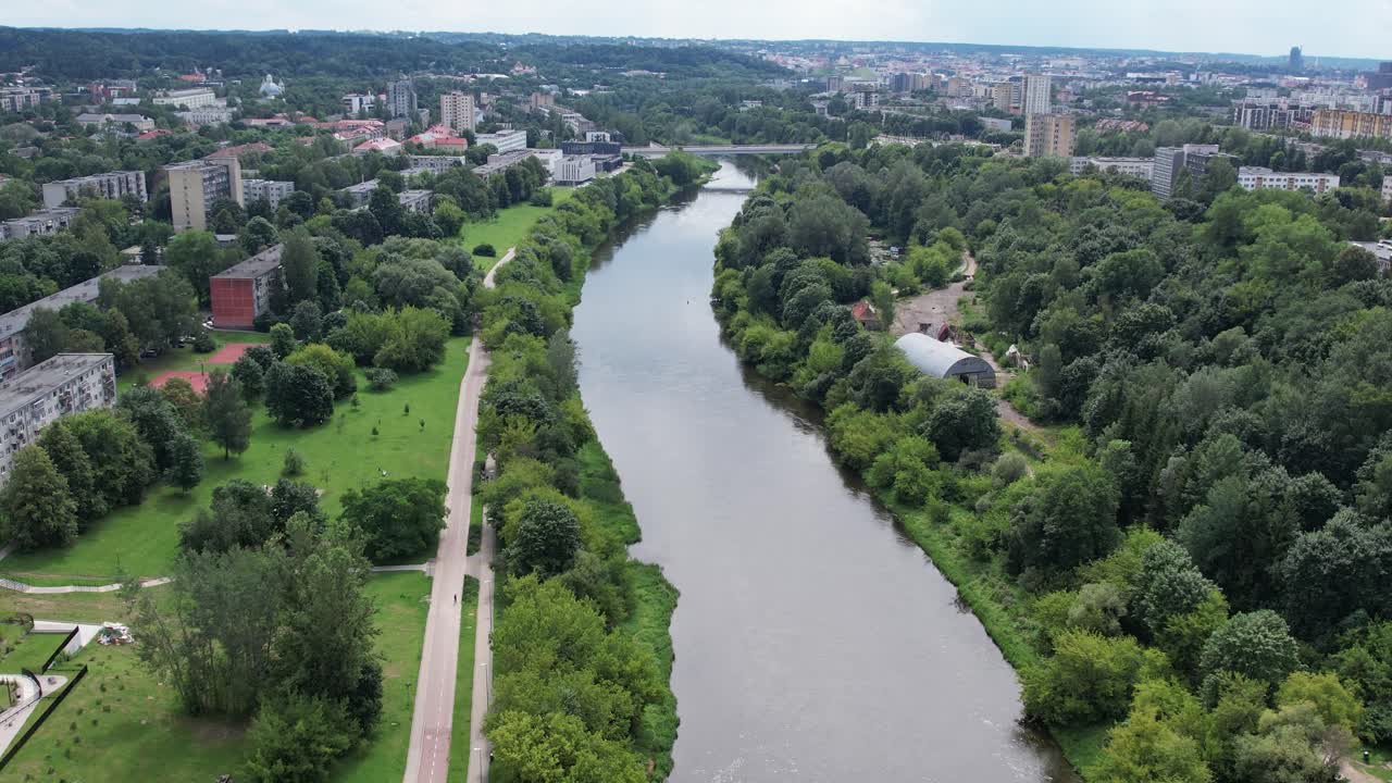 Aerial summer shot of the Neris River in Vilnius, showing green riverbanks, residential districts and walking paths stretching toward the city skyline