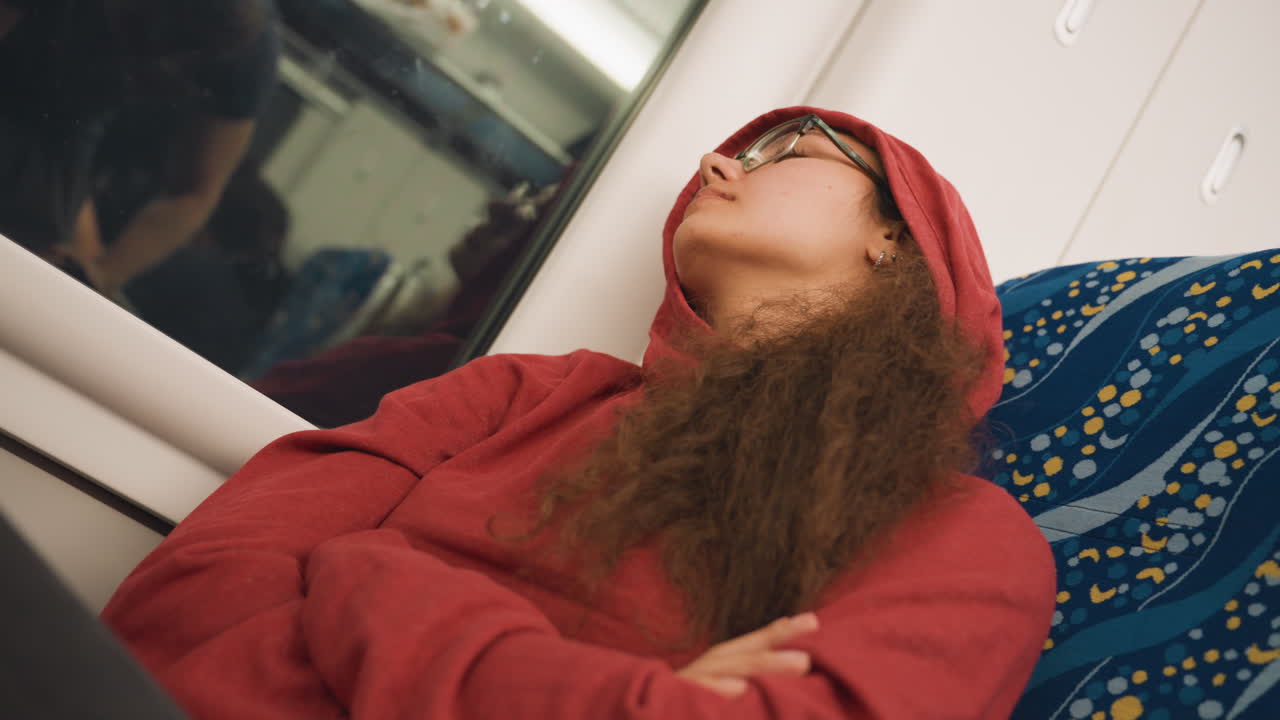 Ung kvinde sover ved togvinduet, universitetsstuderende iført rød hættetrøje og hue, arme krydset over brystet, briller fanger togvognens refleksion, krøllet hår indrammer ansigtet, blåt mønstret sæde.