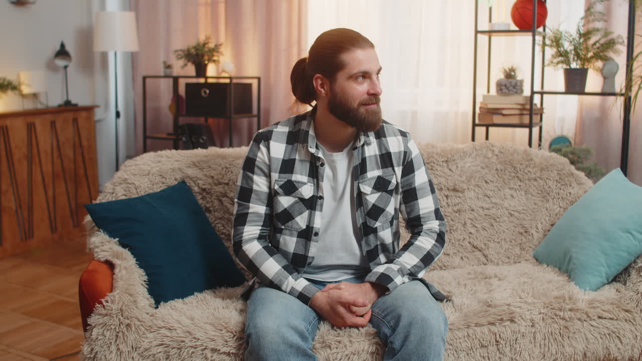 Cheerful young man greeting sits on home sofa wave hey you hello inviting optimism bright gesture